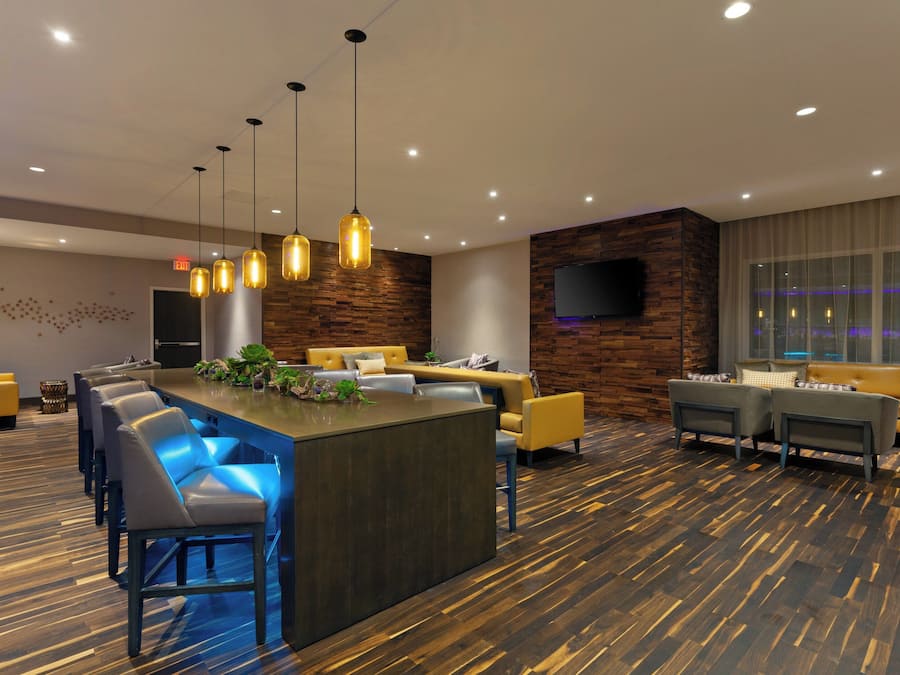 A modern hotel lobby with a central bar area, seating arrangements, and a large television screen.