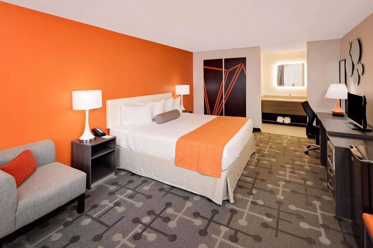 A hotel room with a bed, a desk, a chair, a sofa, and a bathroom visible in the background.