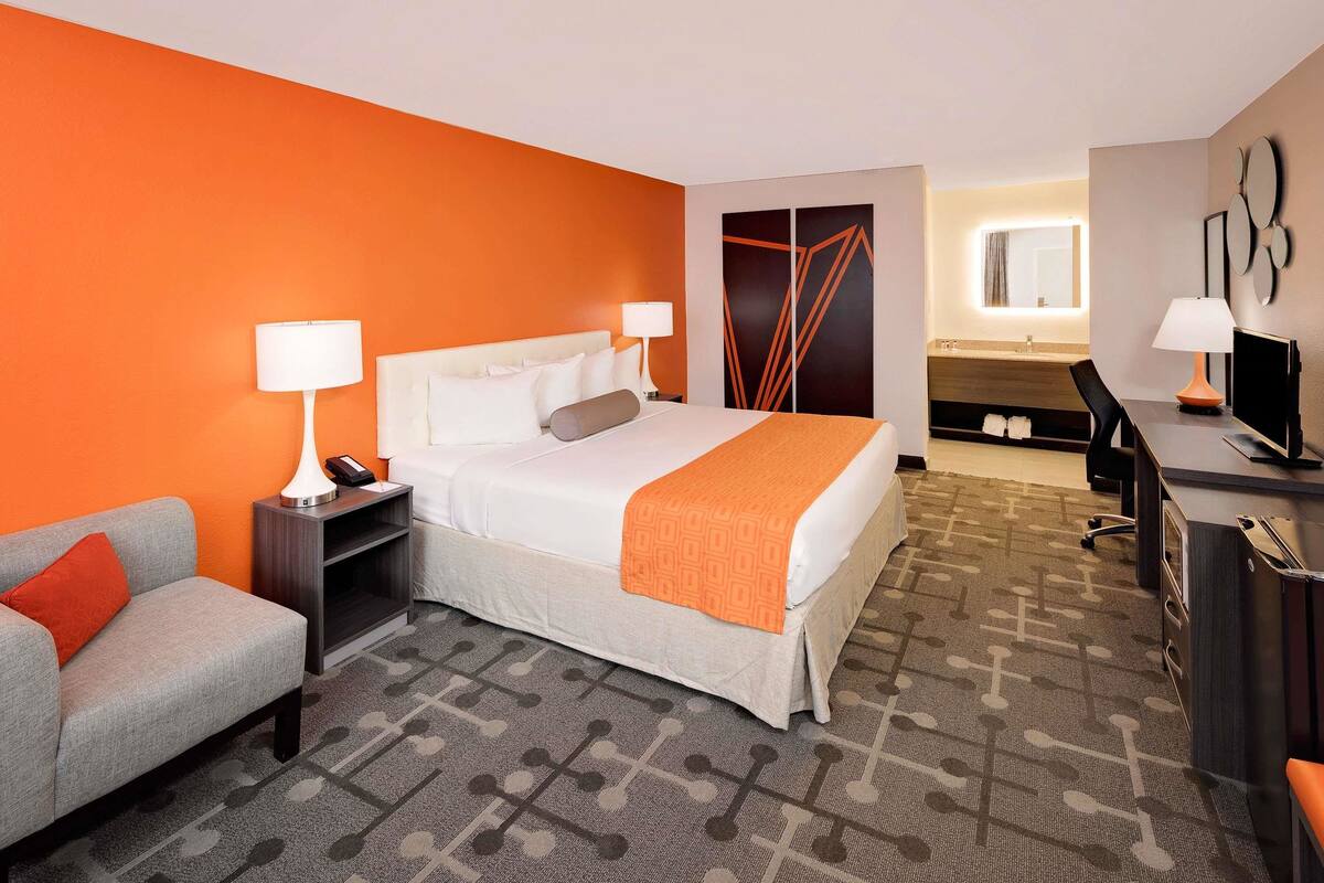 A hotel room with a bed, a desk, a chair, a sofa, and a bathroom visible in the background.