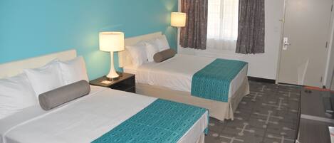 A hotel room with two beds, a desk, a lamp, and a window with curtains.