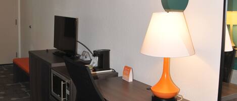 A hotel room with a desk, a computer, a lamp, and a mirror.