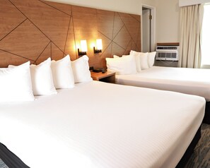 A hotel room with two beds, a wooden headboard, wall-mounted lights, a bedside table, and a window with curtains.