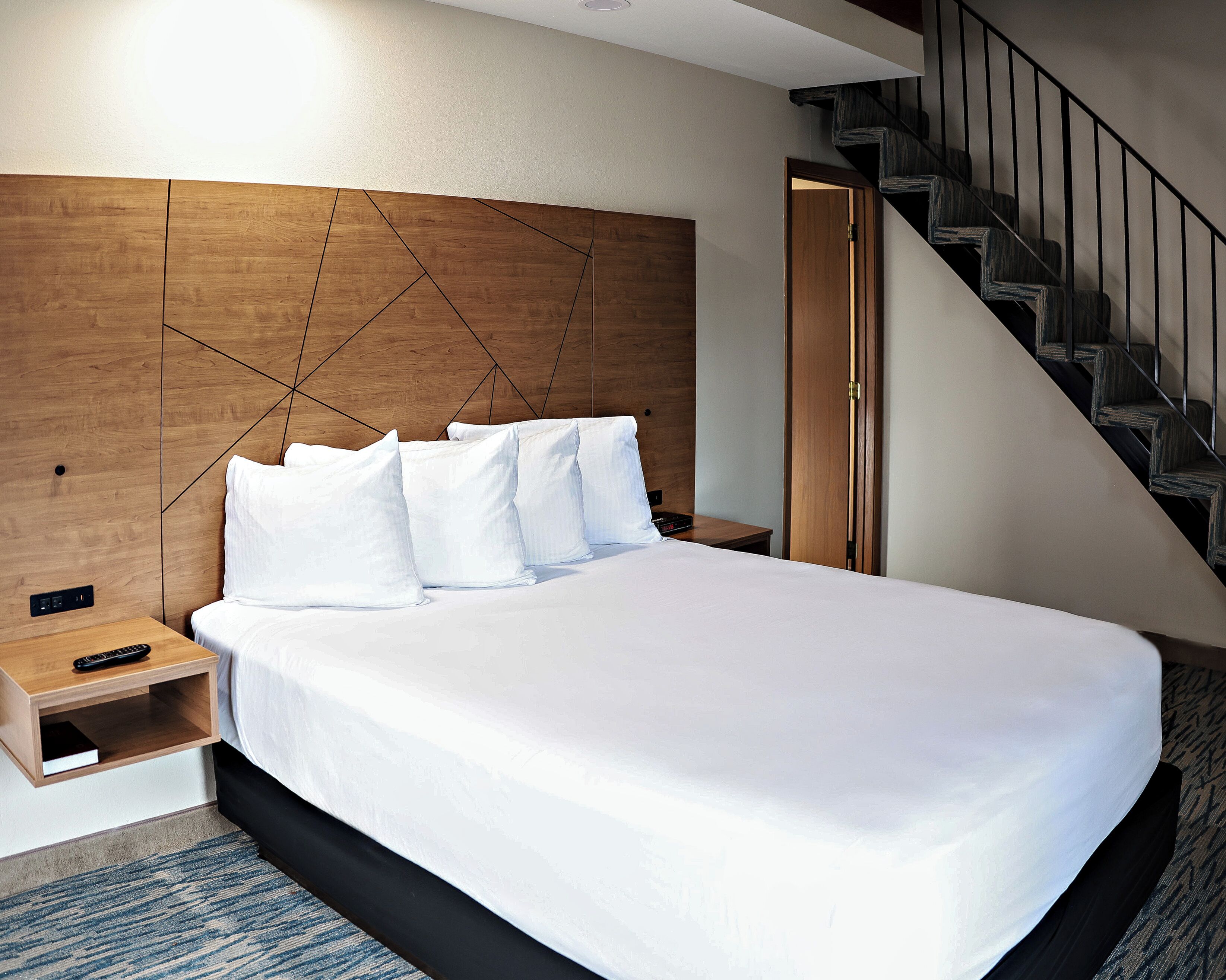 A hotel room with a large bed, a wooden headboard, a nightstand, a remote control, and a staircase.