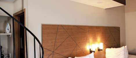 Una habitación de hotel con cabecera de madera, dos camas con ropa blanca, una mesita de noche con lámpara y una escalera con barandilla.