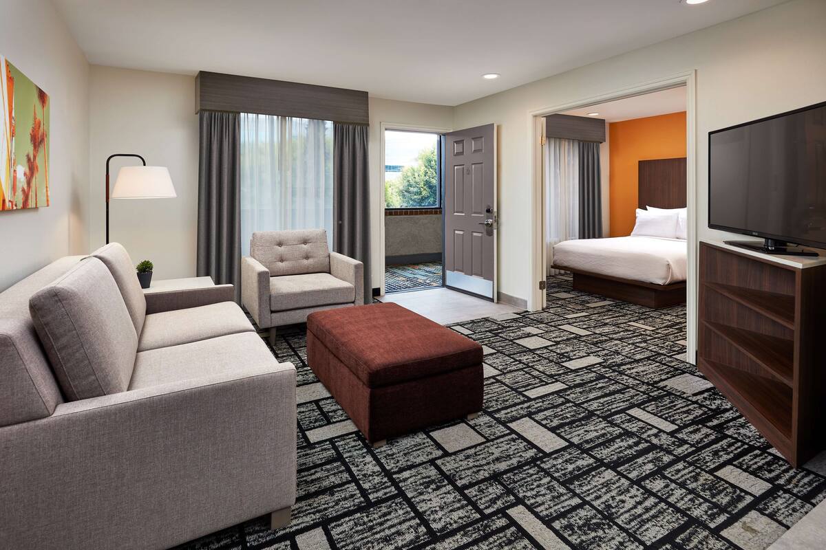 A hotel room with a sofa, armchair, ottoman, a flat-screen TV, and a bed.