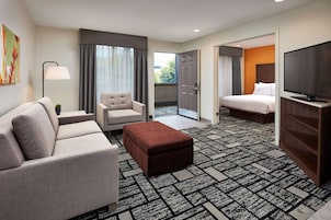 A hotel room with a sofa, armchair, ottoman, a flat-screen TV, and a bed.