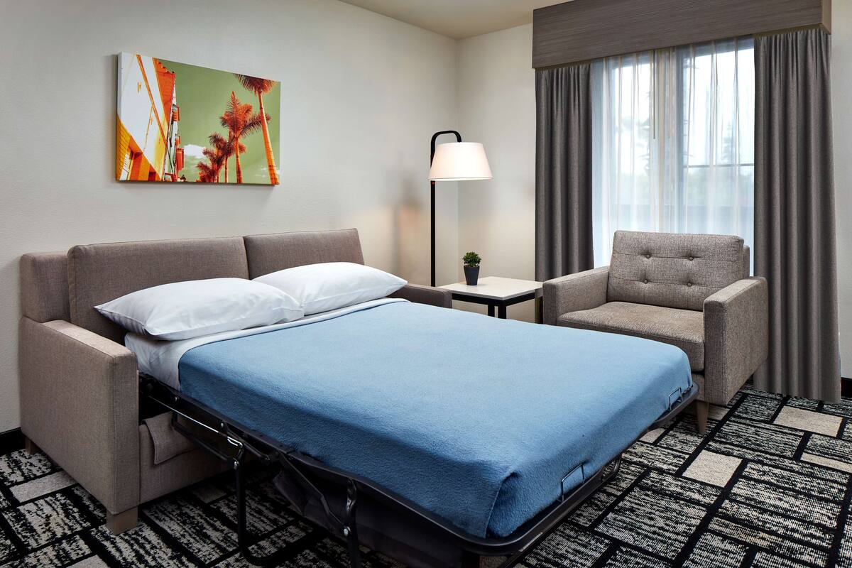 A hotel room with a bed, a sofa, a chair, a lamp, and a painting on the wall.