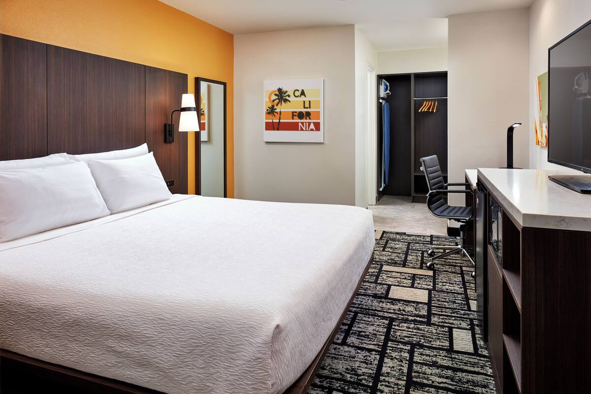 A hotel room with a large bed, a desk with a chair, a TV, a wardrobe, and a wall with a tropical-themed artwork.