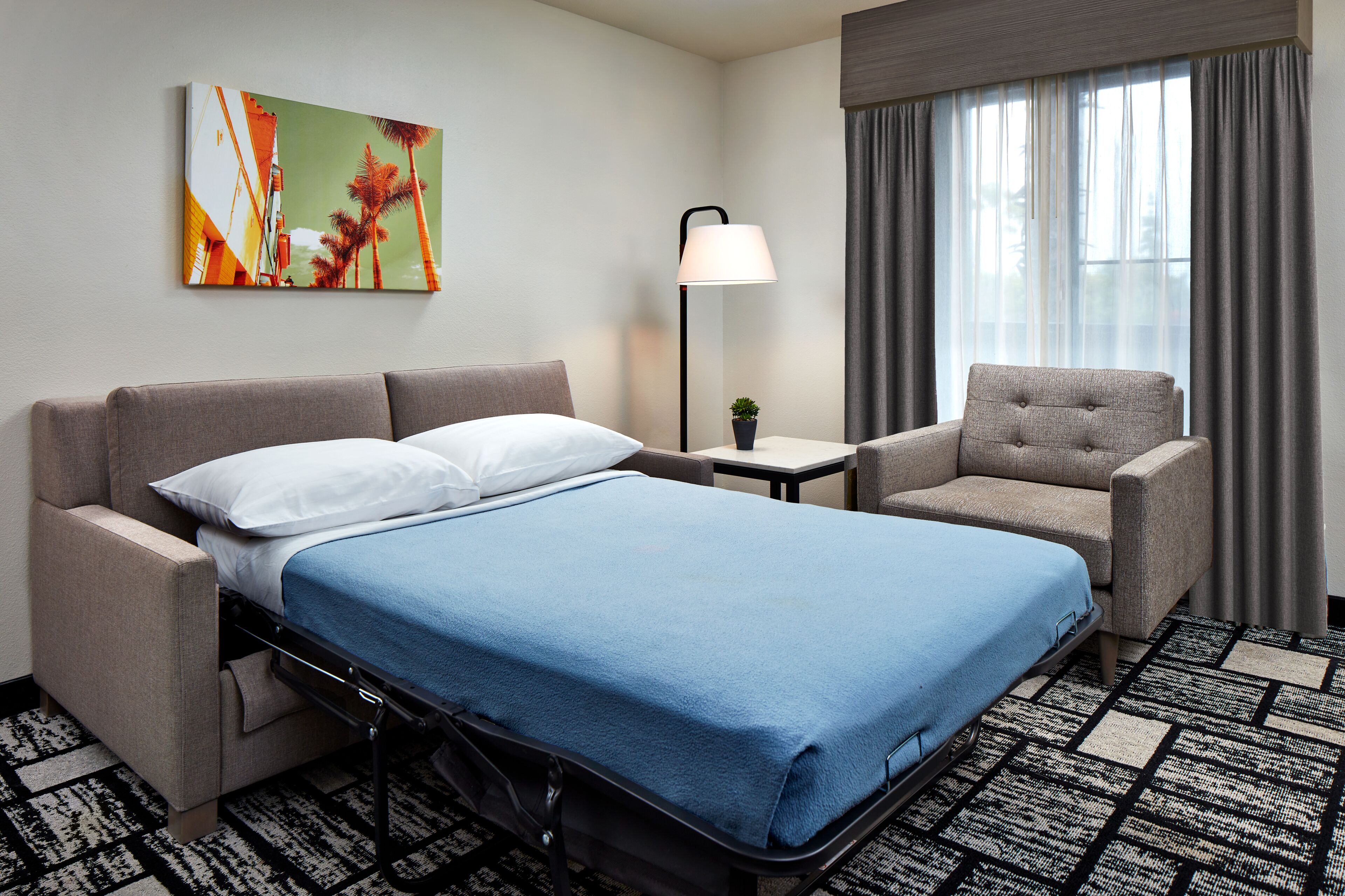 Hotelkamer met een bed, een bank, een stoel, een lamp, een tafeltje en een raam met gordijnen.
