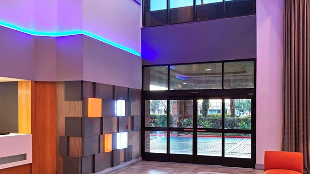A modern lobby with a large glass window, a reception desk, and a seating area with an ottoman and chairs. - Best Western Plus Meridian Inn & Suites, Anaheim-Orange (Orange)