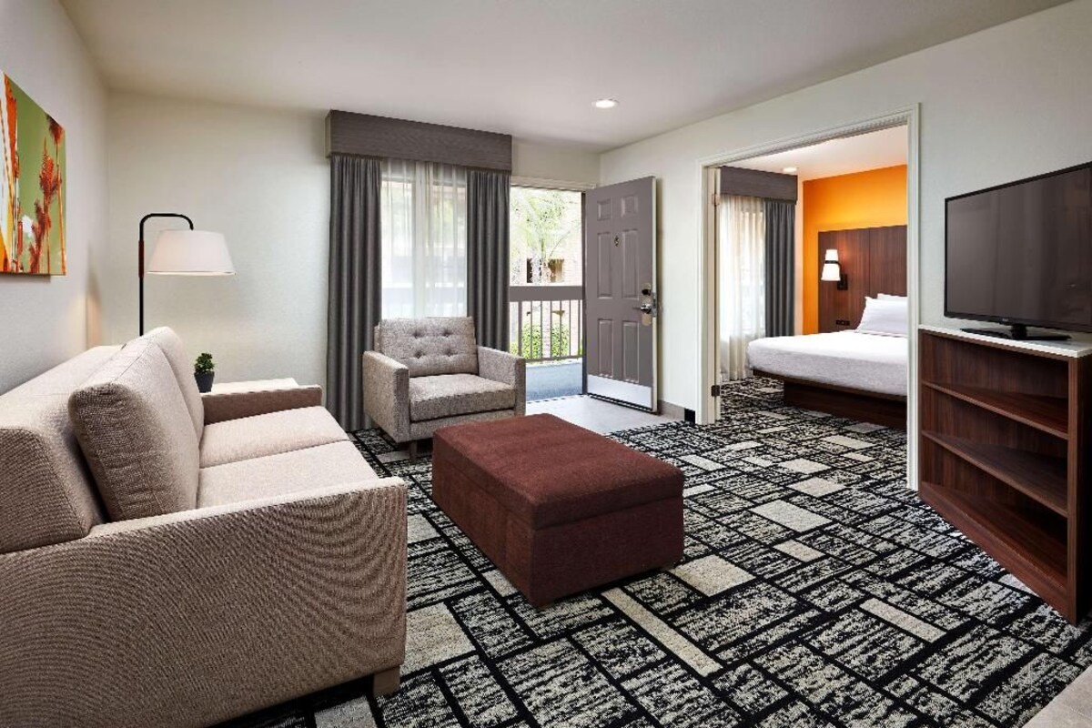Habitación de hotel con sofá, sillón, reposapiés, televisión y alfombra estampada.
