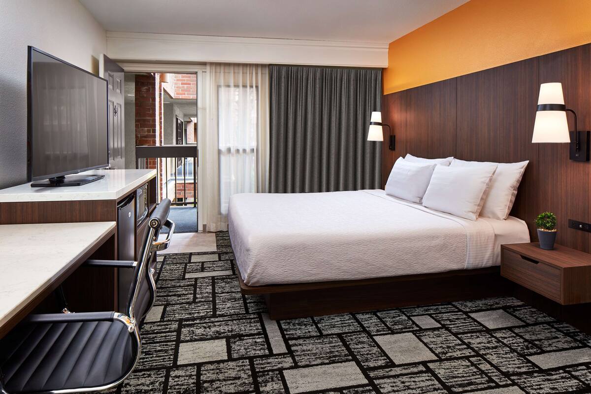 A hotel room with a bed, desk, chair, TV, and a small plant.