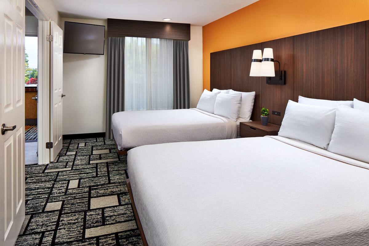A hotel room with two beds, a TV mounted on the wall, a nightstand with a lamp, and a patterned carpet.