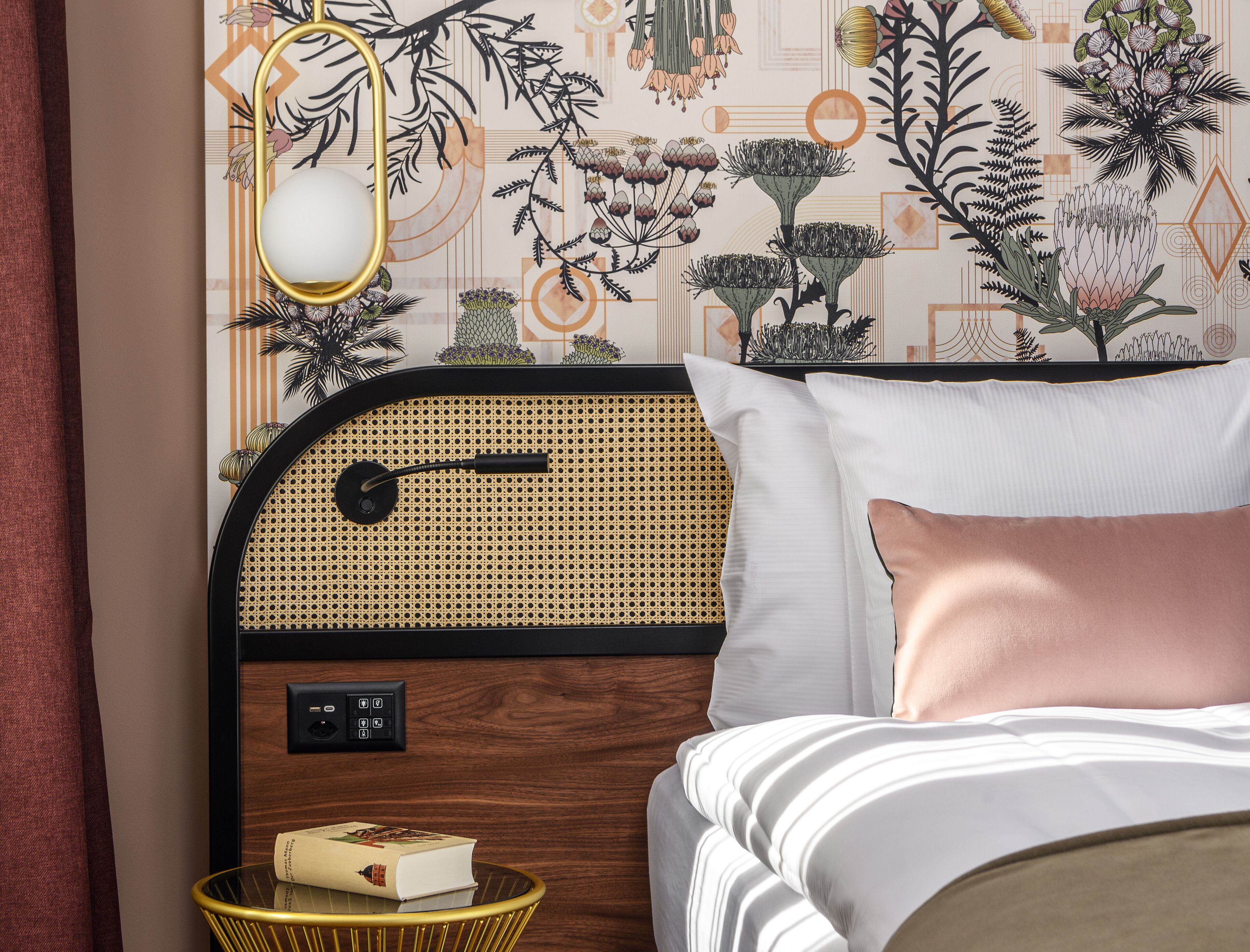 A bedroom with a rattan headboard, a bedside table with a book, and a decorative wall with plant illustrations.