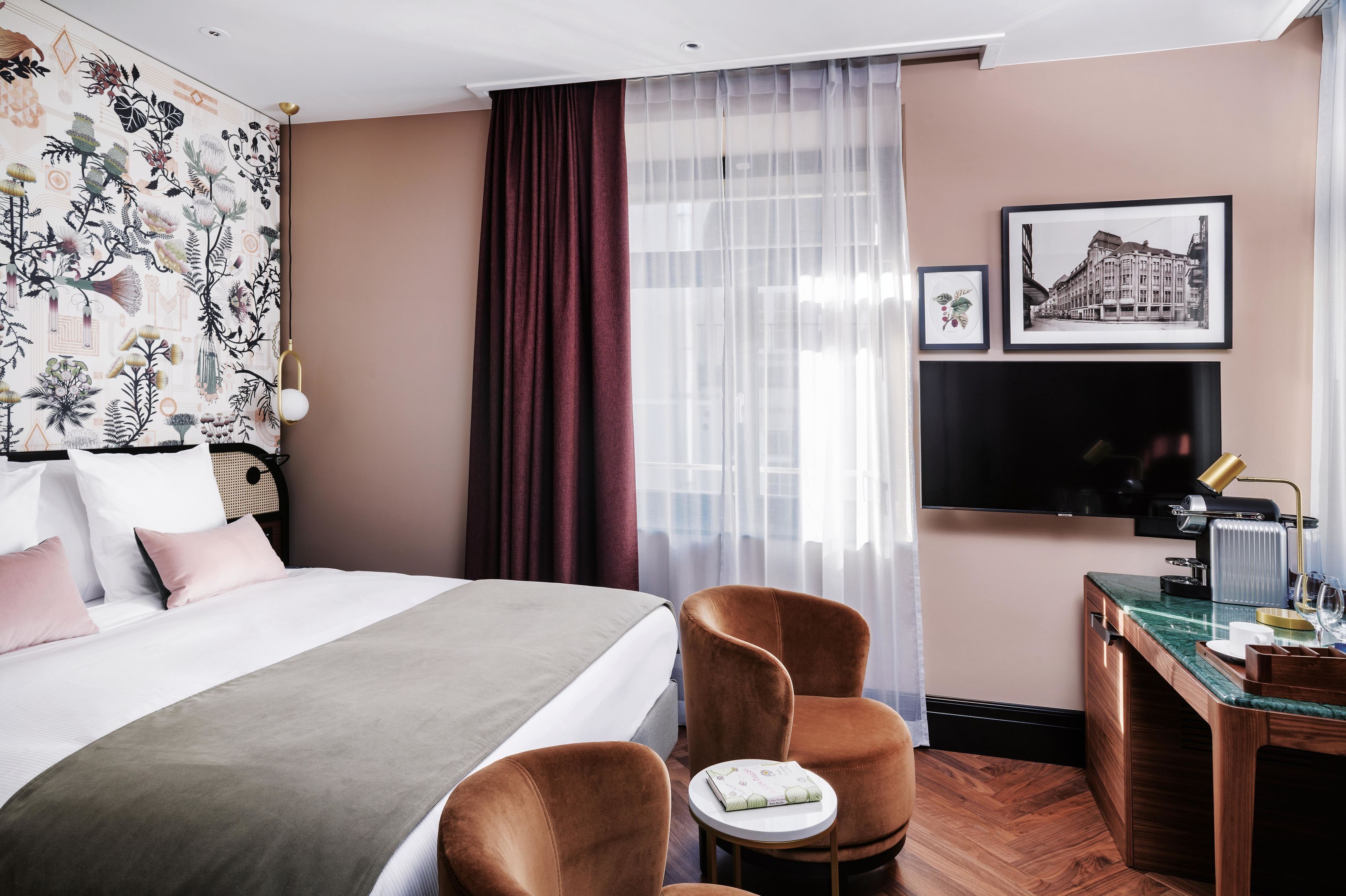 A hotel room with a bed, two chairs, a desk, a TV, and a window with sheer curtains.