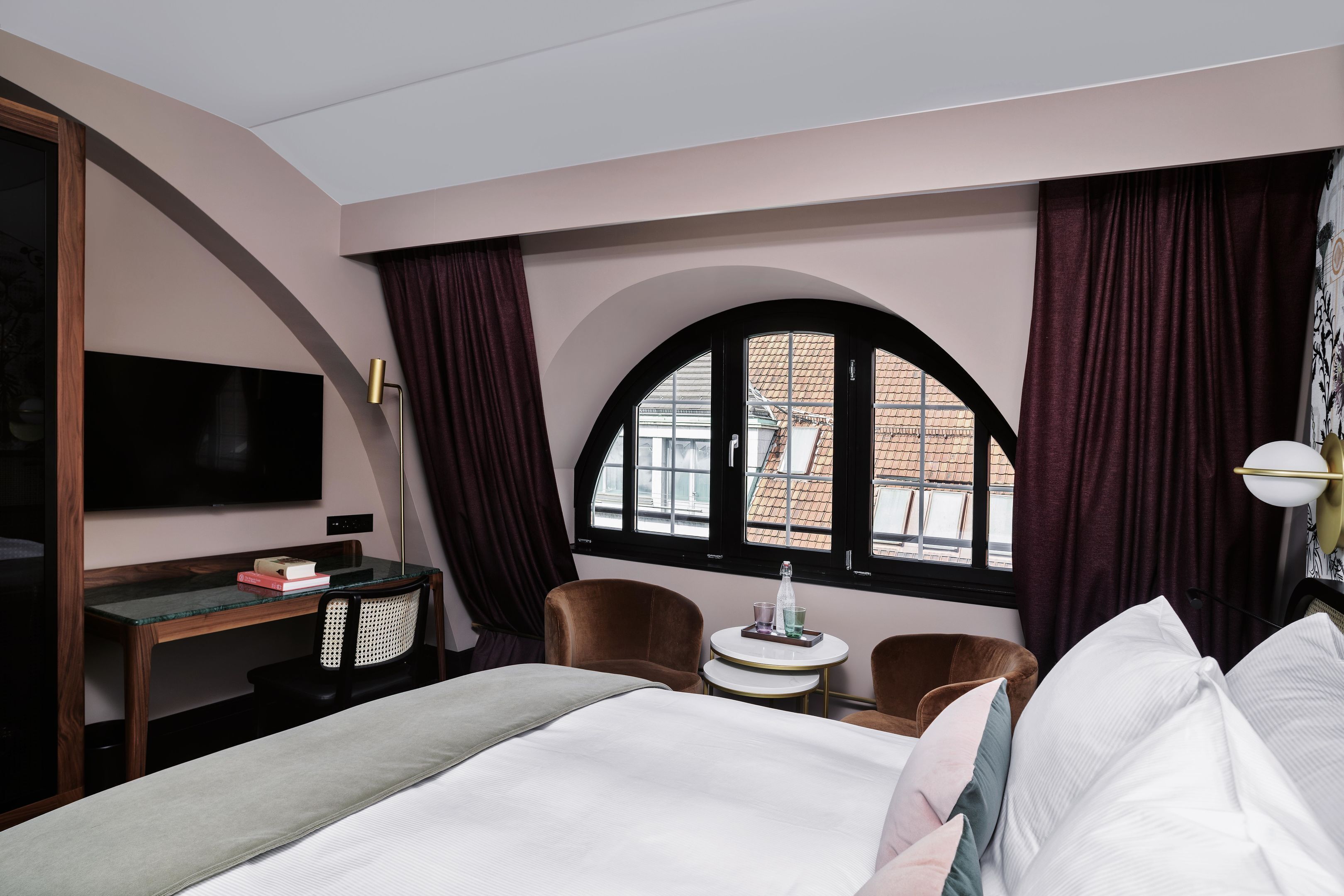 Habitación de hotel con cama, escritorio, silla, mesita, televisor y ventana con cortinas.