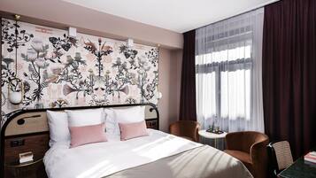 A hotel room with a bed, a chair, a small table, and a window with curtains.