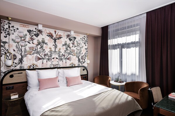 A hotel room with a bed, a chair, a small table, and a window with curtains.