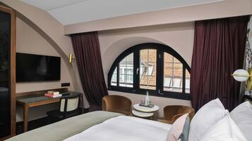 Ein Hotelzimmer mit Bett, Schreibtisch, Fernseher und einem Fenster mit Vorhängen sowie Blick auf Gebäude.
