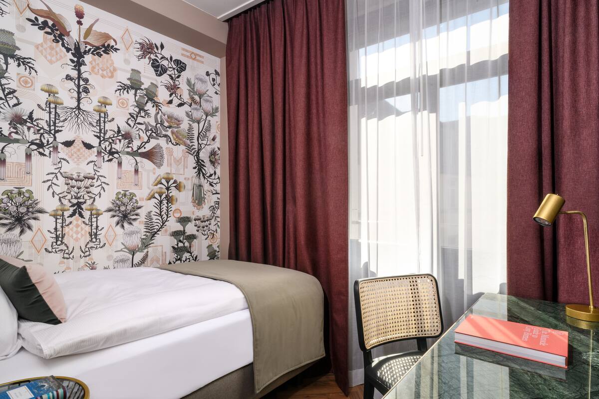 Une chambre d’hôtel avec un lit, une chaise, un bureau avec une lampe et une fenêtre avec des rideaux.