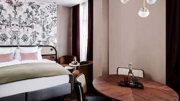 Et hotelværelse med en seng, et rundt spisebord, en stol, et lille bord med et tepotteskjul og en væg med et blomstret gobelin.