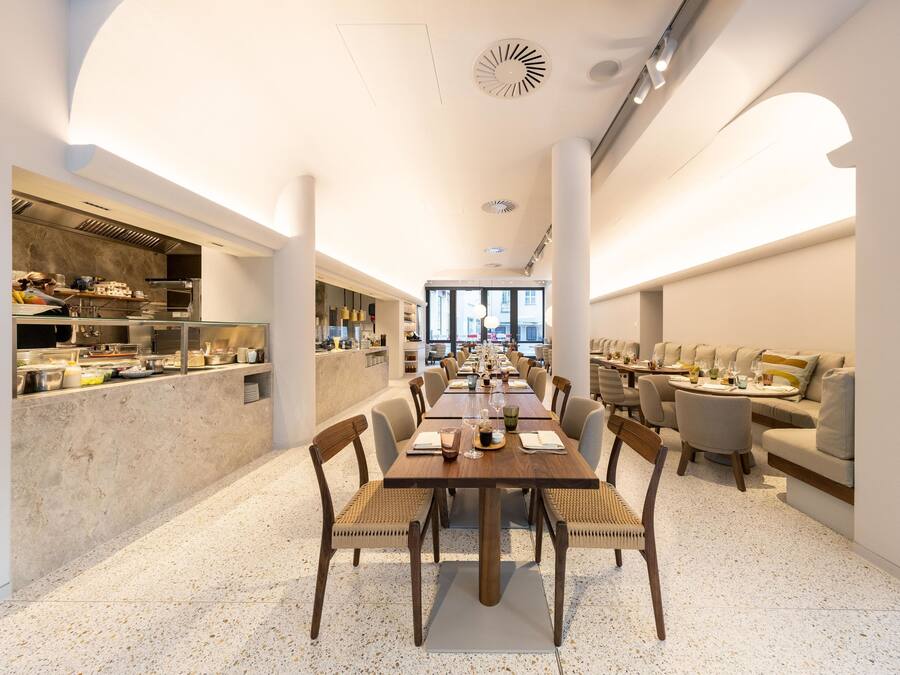A modern dining area with a long wooden table, cushioned chairs, and a kitchen counter on one side.