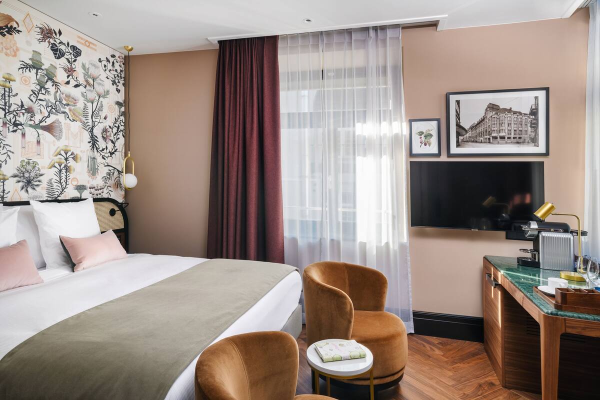 Une chambre d’hôtel comprenant un lit, deux chaises, une petite table, un bureau, un miroir et une fenêtre avec des rideaux légers.