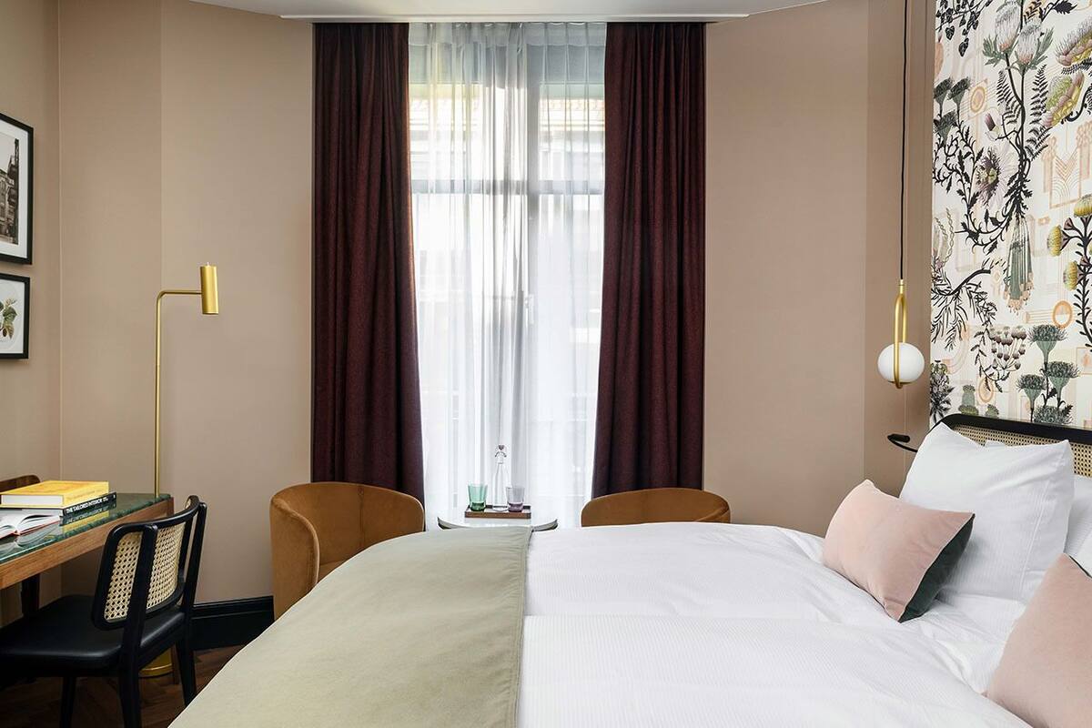 Une chambre d’hôtel avec un lit, deux chaises, un bureau avec une lampe et une fenêtre avec des rideaux.
