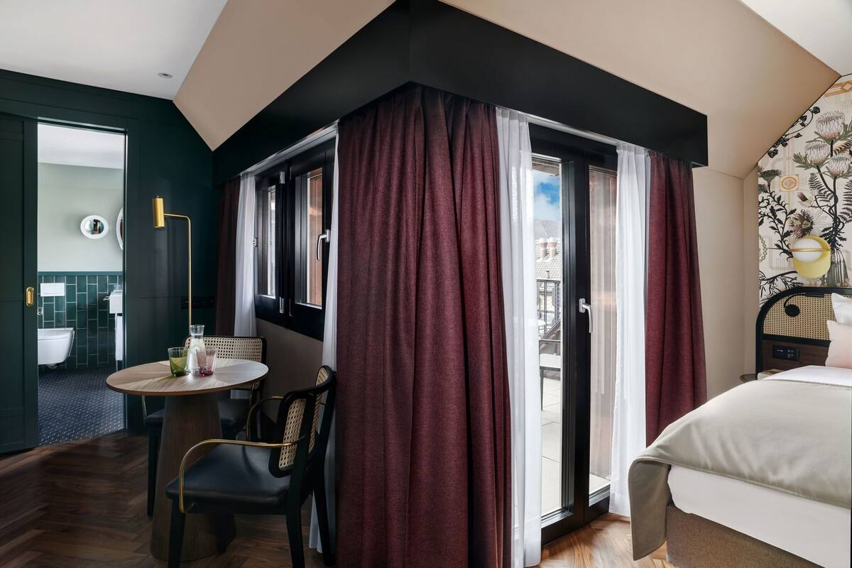 Une chambre d’hôtel avec un lit, une table ronde entourée de chaises, une salle de bain visible à travers une porte ouverte et un balcon avec vue.
