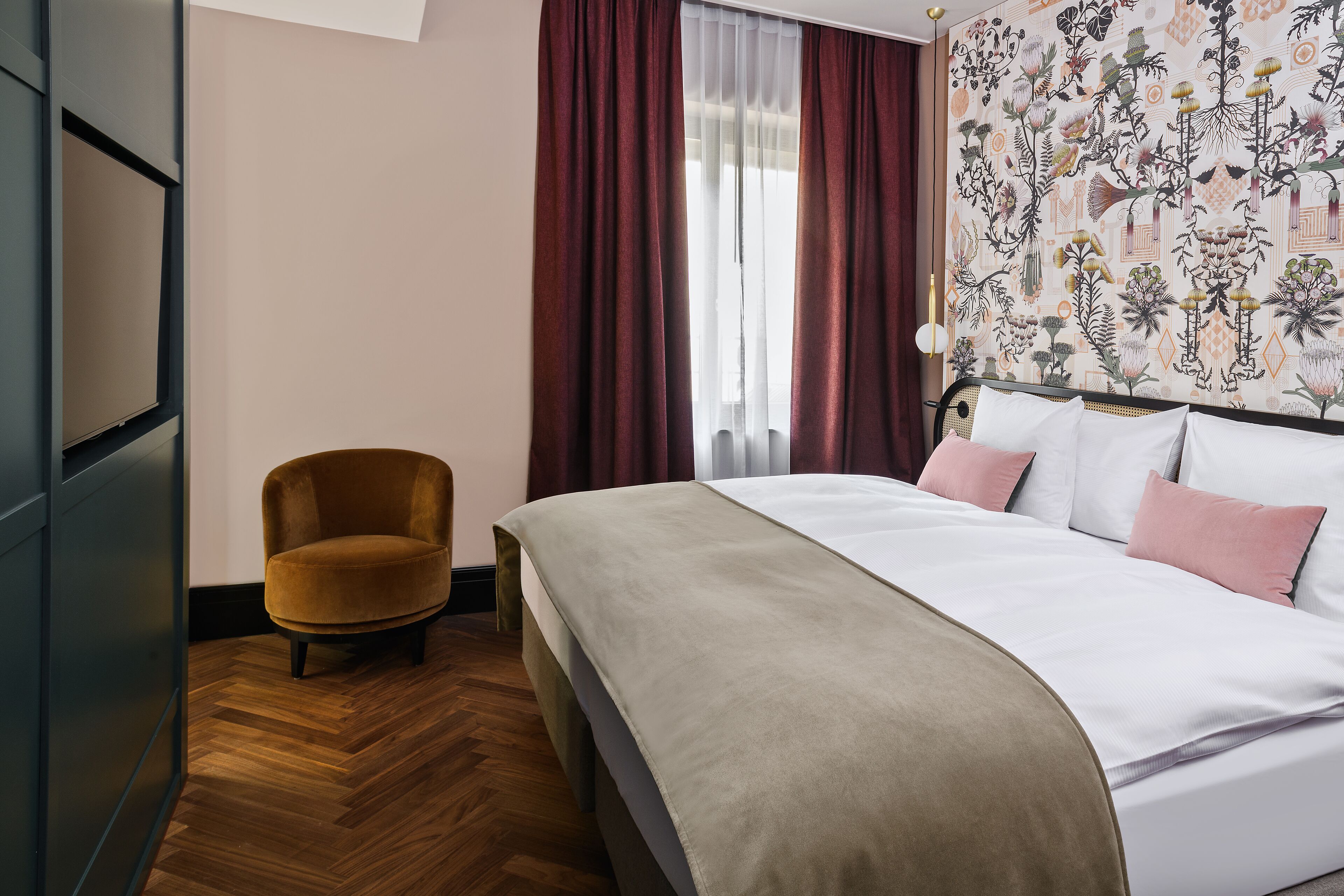 A hotel room with a bed, a chair, a wooden floor, and a floral wallpaper.
