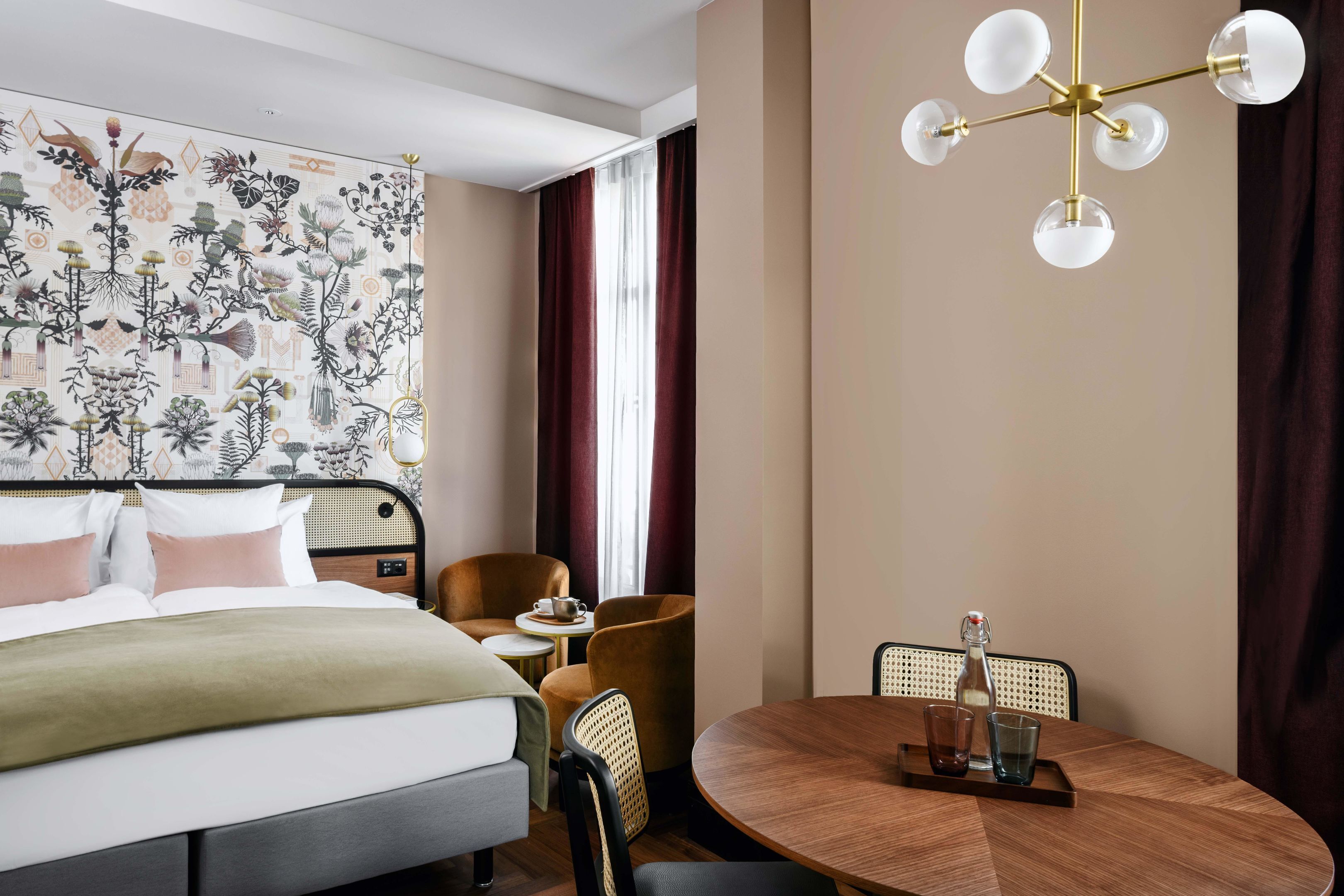 A hotel room with a bed, a round dining table, a chandelier, and a wall with a botanical print.