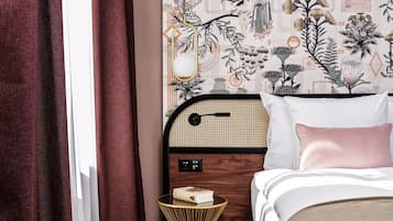 Habitación de hotel con cama, mesita de noche con un libro, espejo en la pared y pared decorativa con motivo geométrico.
