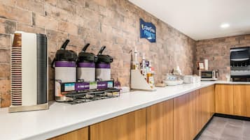 A kitchen area with a brick wall, a coffee station with branded dispensers, a microwave, and a flat-screen TV.