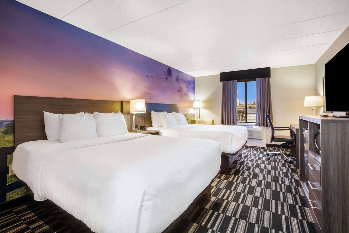 Habitación de hotel con dos camas, un escritorio, un televisor y un ventanal con cortinas.