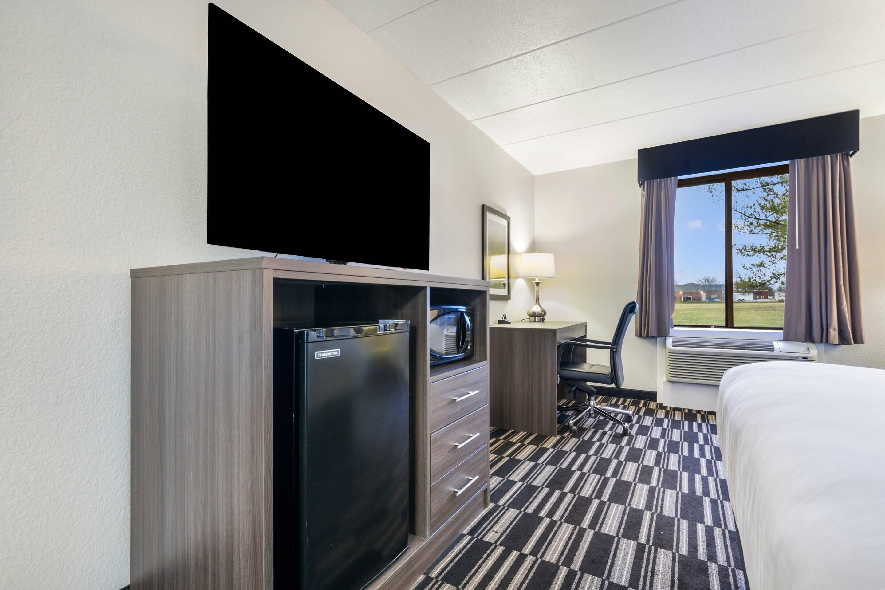 A hotel room with a bed, a desk with a chair, a television mounted on the wall, a refrigerator, a microwave, a lamp, and a window with curtains.