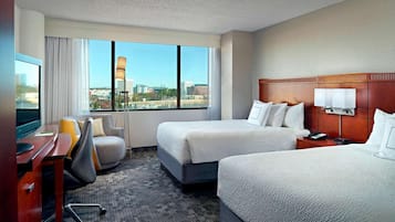 A hotel room with two beds, a desk with a computer, a chair, and a large window offering a city view.
