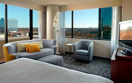 A hotel room with a large bed, a grey sofa with yellow pillows, a grey armchair, a small table, and a TV.