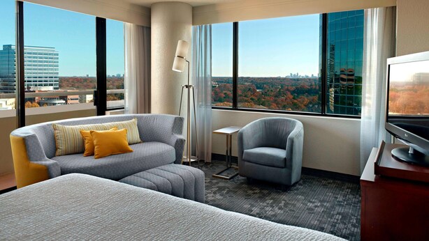 A hotel room with a large bed, a grey sofa with yellow pillows, a grey armchair, a small table, and a TV.