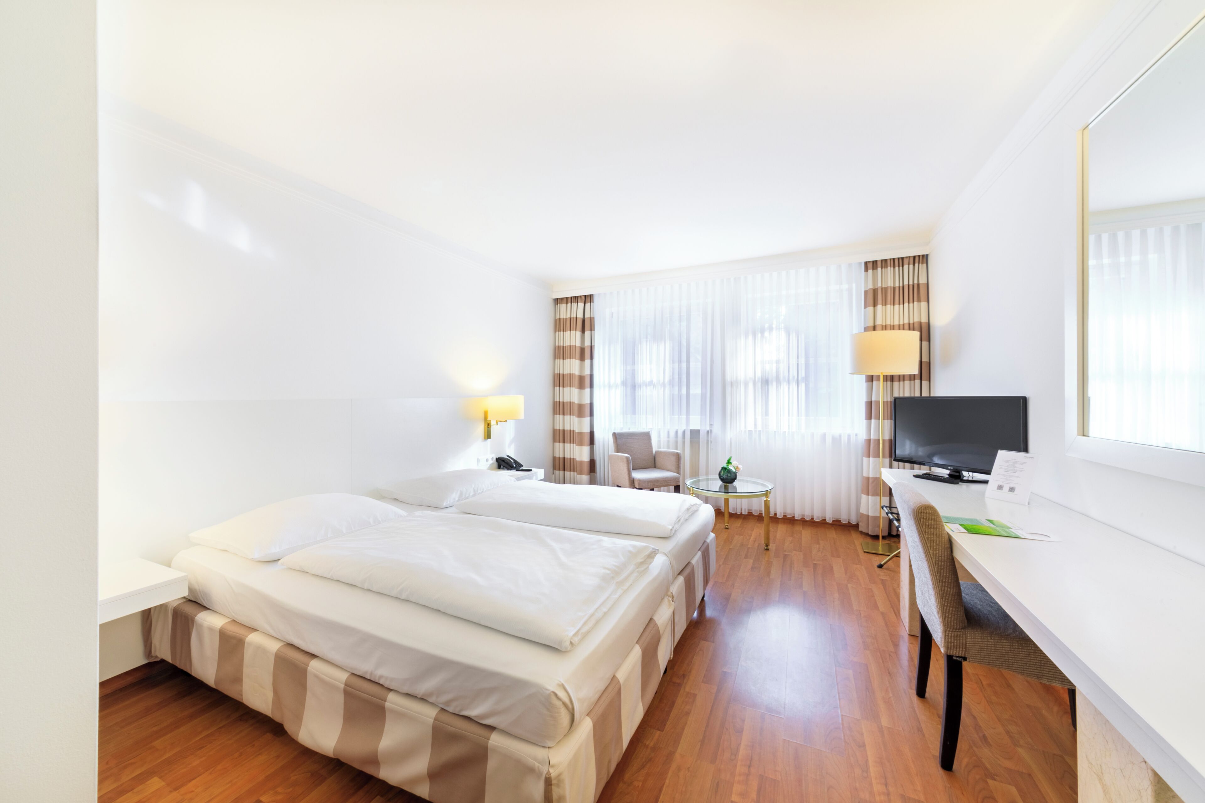 Quarto de hotel com duas camas, uma escrivaninha, televisão e janela com cortinas.