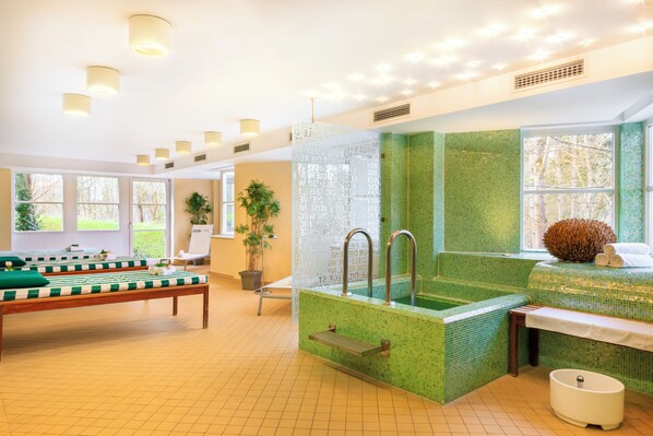 Une salle de spa avec un mur recouvert d’une mosaïque verte, un jacuzzi et une table de massage.