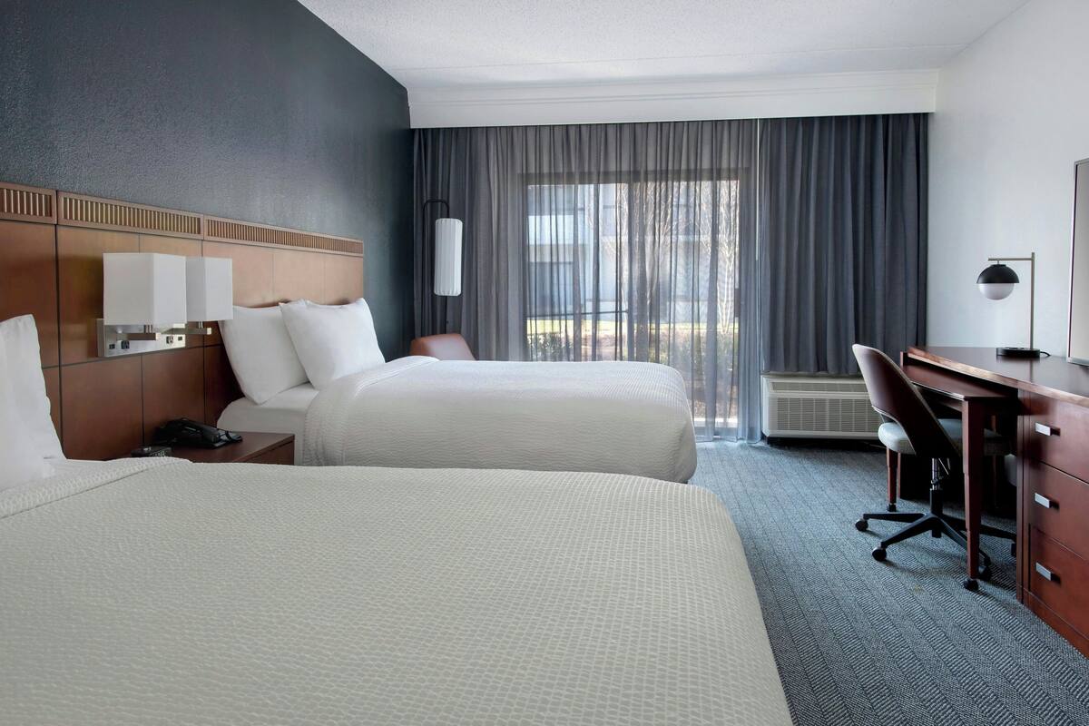 Habitación de hotel con dos camas, un escritorio, una silla, una lámpara y un ventanal con cortinas.