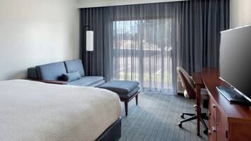 A hotel room with a large bed, a desk, a chair, a sofa, and a view of the outdoors through the window.