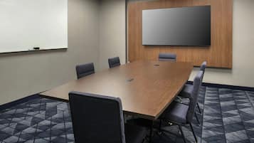A conference room with a large wooden table, six chairs, a whiteboard, and a large screen on the wall.