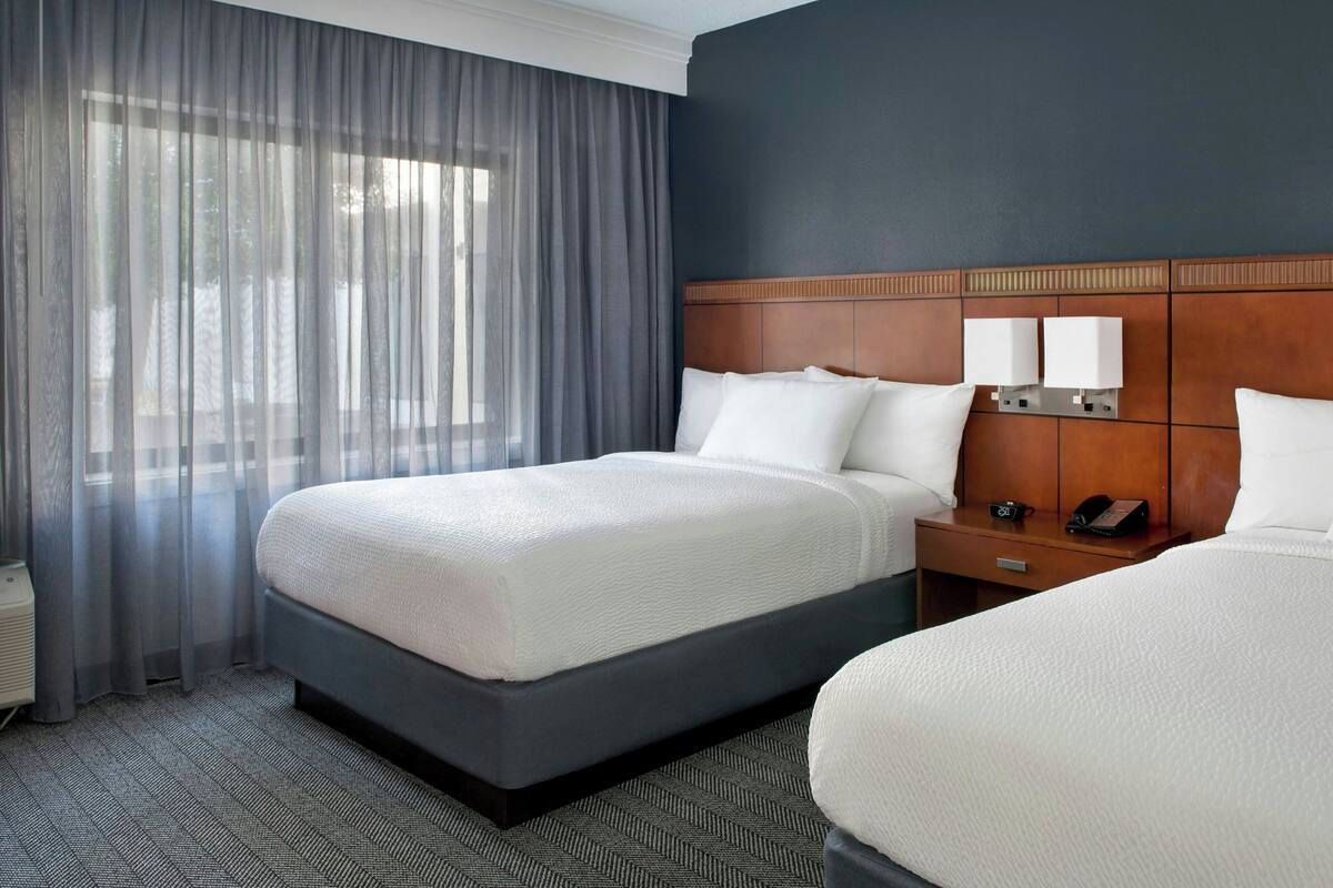 Habitación de hotel con dos camas, cabecero de madera, mesita de noche con teléfono y ventana con cortinas translúcidas.