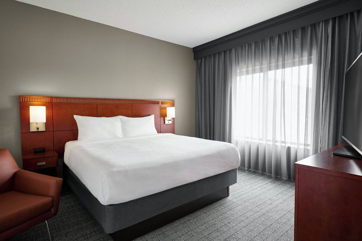 Ein Hotelzimmer mit einem großen Bett, einem Holzkopfende, zwei Nachttischlampen, einem Fernseher auf einem hölzernen Standfuß, einem roten Sessel und einem großen Fenster mit durchsichtigen Vorhängen.