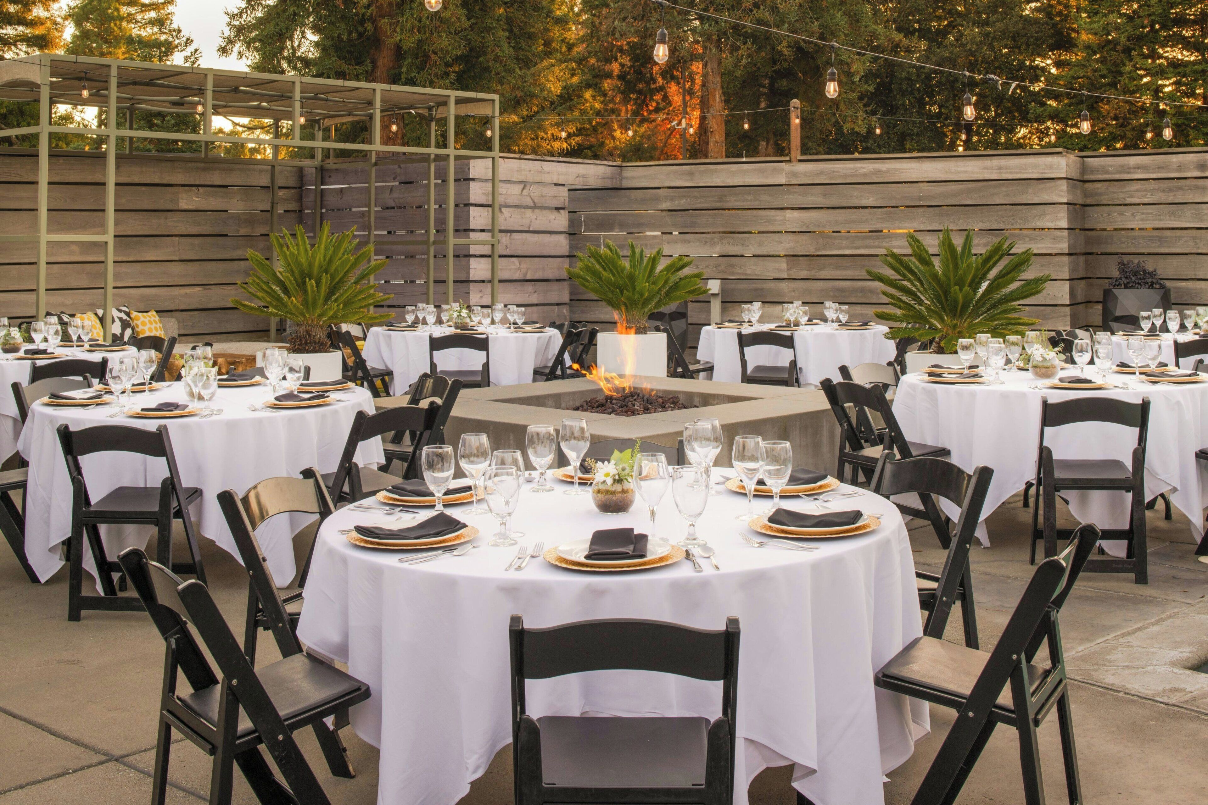 An outdoor dining area with round tables set for a meal, surrounded by chairs and potted plants.