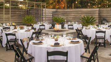 An outdoor dining area with round tables set for a meal, surrounded by chairs and potted plants.