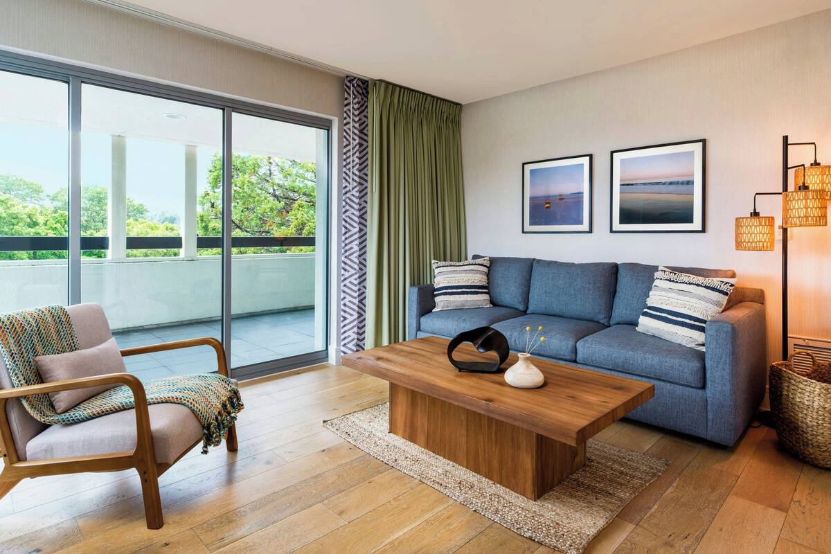 A modern living room with a sofa, armchair, wooden coffee table, and large windows.