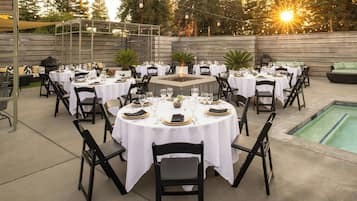 Un área de comedor al aire libre con mesas redondas dispuestas para una comida, rodeada de sillas y una piscina al fondo.
