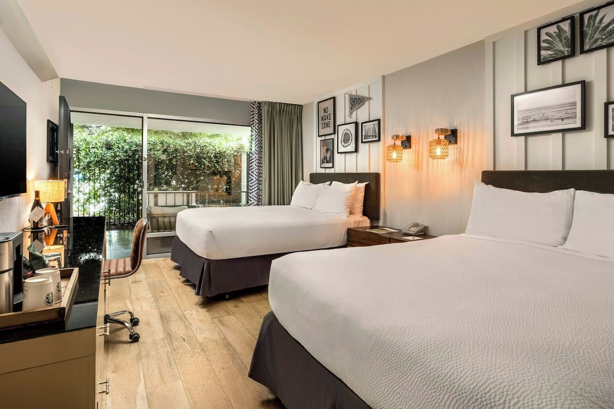 A hotel room with two beds, a desk with a lamp, a TV, and a view of greenery through the window.